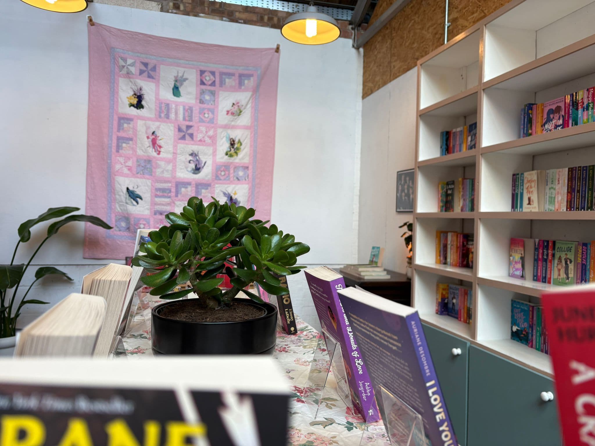 Interior of Cupid's Bow Books showing shelves of romance novels, books displayed on a floral tablecloth, a jade plant, and a handmade fairy quilt hanging on the wall